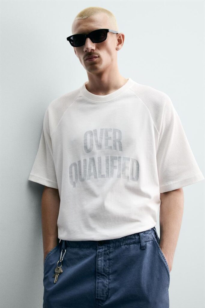 White t-shirt featuring oversized grey “OVER QUALIFIED” typography graphic on front placement.
